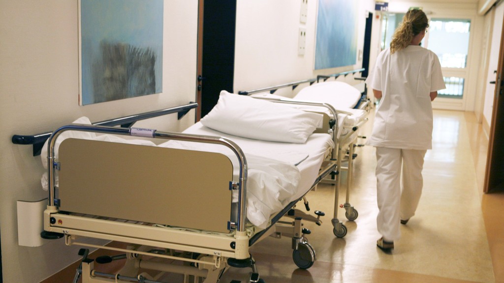 Eine Krankenschwester geht auf einem Flur an einem leeren Bett vorbei.