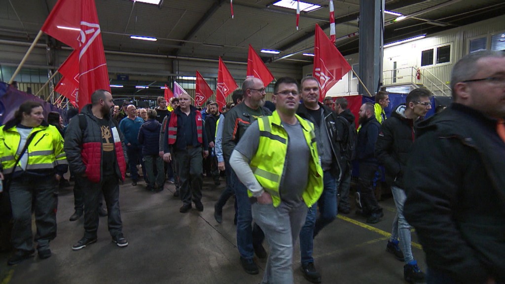 Foto: Menschen mit roten Fahnen und Warnwesten in Fabrikhalle