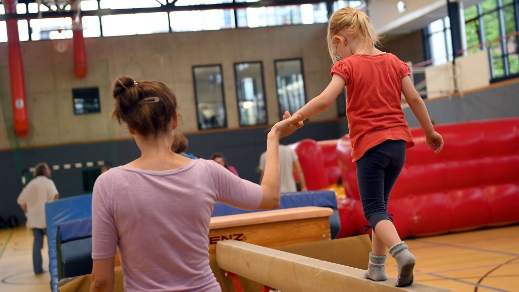 Ein Kind wird beim Kinderturnen von einer Erwachsenen auf dem Schwebebalken geführt