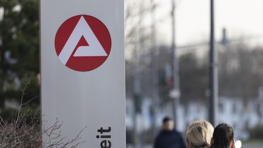 Foto: Stele mit dem Zeichen, Logo der Agentur für Arbeit vor einem Gebäude. Personen gehen daran vorbei. 