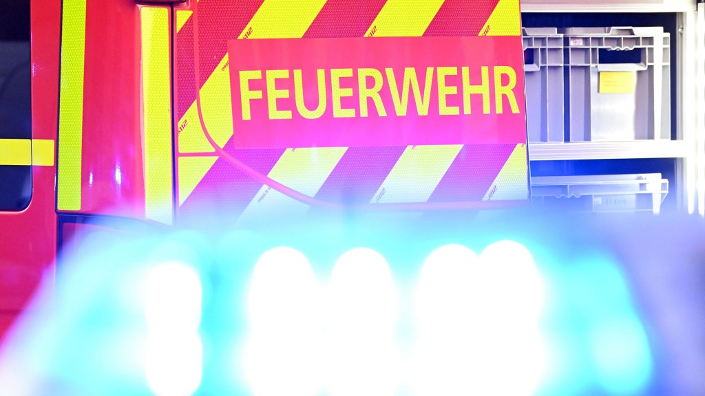 Foto:  Ein Fahrzeug der Feuerwehr steht bei einem Einsatz auf einer Straße, während im Vordergrund das Blaulicht eines Polizeifahrzeugs zu sehen ist