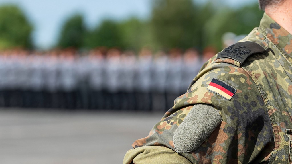 Foto: Ein Soldat der Bundeswehr steht in Flecktarnuniform mit Deutschlandabzeichen auf dem Ärmel und eine Truppe im Hintergrund.