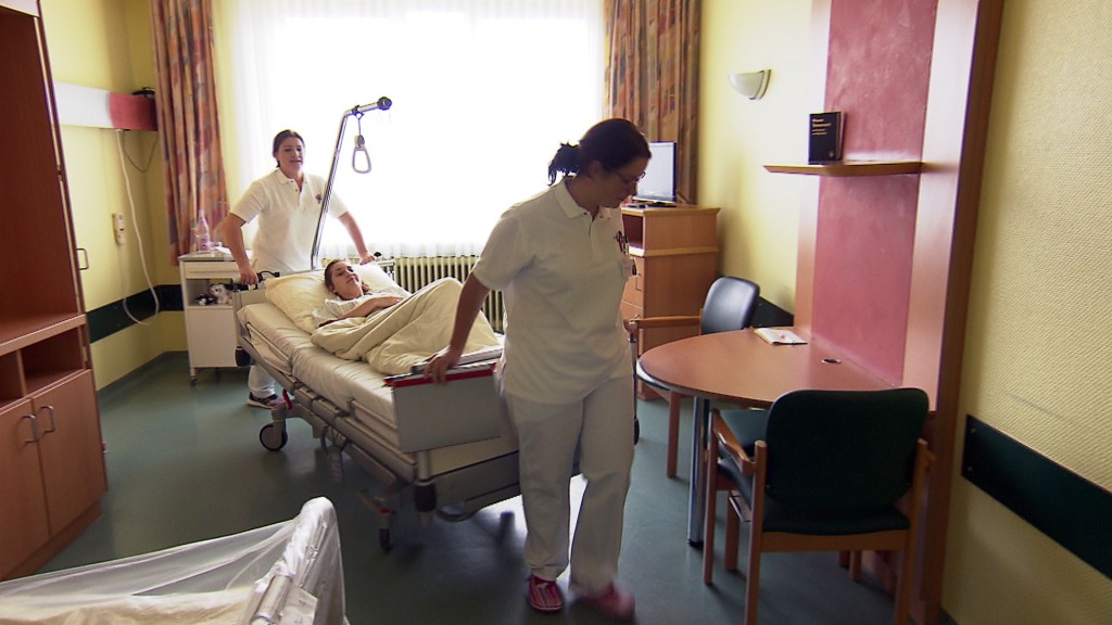 Foto: Zwei Krankenpflegerinnen schieben Krankenbett mit Patienten aus dem Zimmer