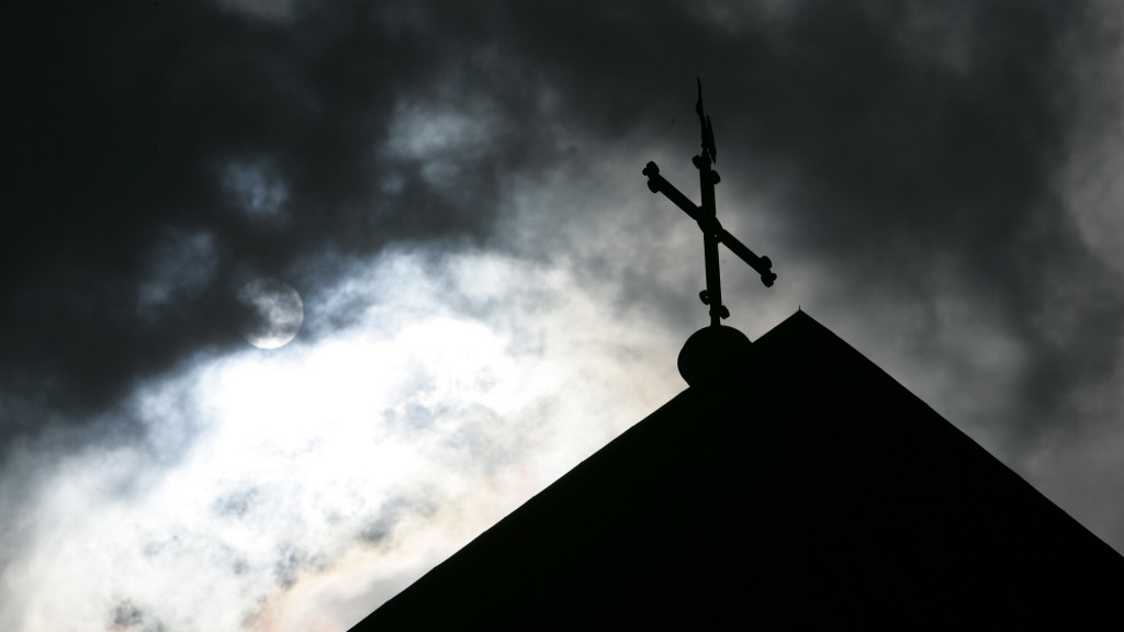 Im Gegenlicht und vor wolkenverhangenem Himmel ist die Kirchturmspitze mit Kreuz zu sehen. (Foto: picture alliance/dpa | Friso Gentsch)