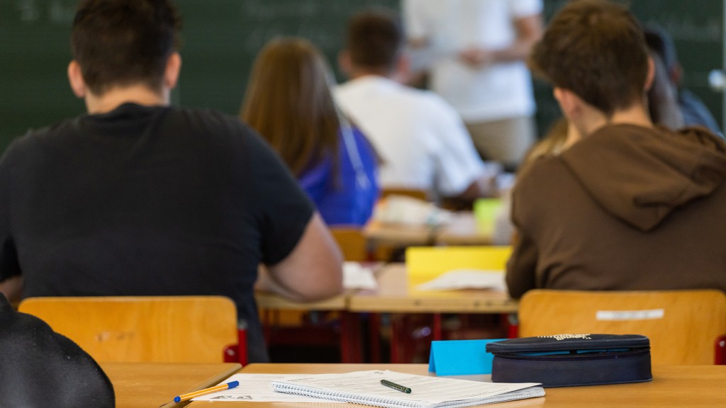 Arbeitsmaterialien liegen auf einem Tisch in einem Klassenzimmer während dahinter Schüler und ihr Lehrer zu sehen sind.