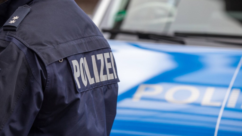 Foto: Schriftzug Polizei auf der Rückseite einer Jacke eines Polizisten vor einem Streifenwagen 