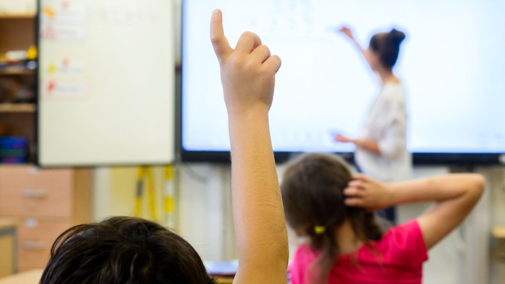 Ein Schüler meldet sich per Handzeichen während eine Lehrerin auf eine digitale Schultafel im Klassenraum einer 4. Klasse einer Grundschule schreibt