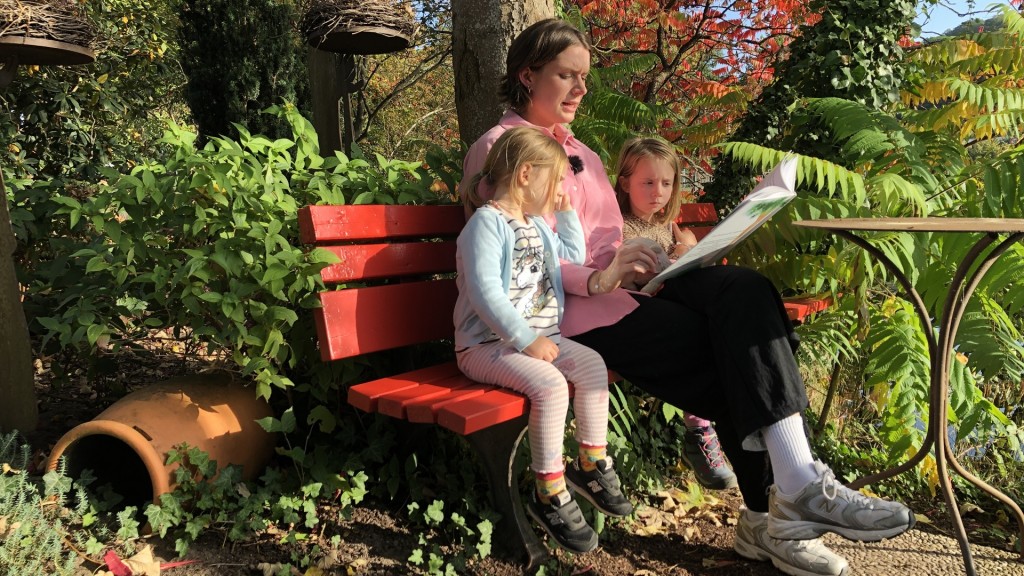 Foto: Eine Frau sitzt mit zwei Kindern auf einer Bank und liest ihnen aus dem Buch 
