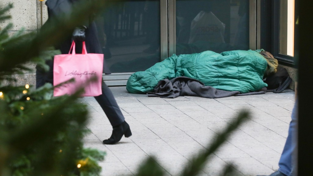 Passanten gehen mit Einkaufstüten in einer Fußgängerzone in der Innenstadt an einem schlafenden Obdachlosen vorbei.