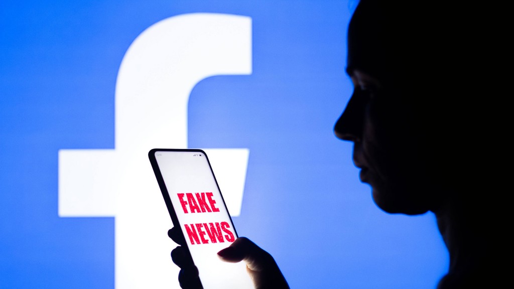 Eine Frau hält ein Smartphone mit der Aufschrift „Fake News“ vor dem Facebook-Logo hin der Hand