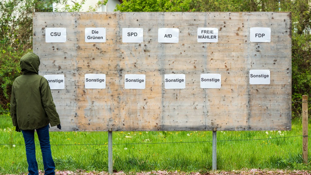 Eine Person mit Kapuzenjacke steht vor einer leeren Plakatwand mit Beschriftung verschiedener Parteien