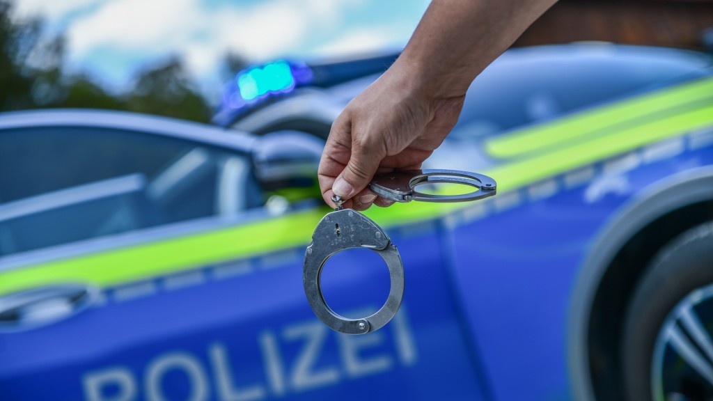 Eine Person hält Handschellen in der Hand. Im Hintergrund steht ein Polizeifahrzeug