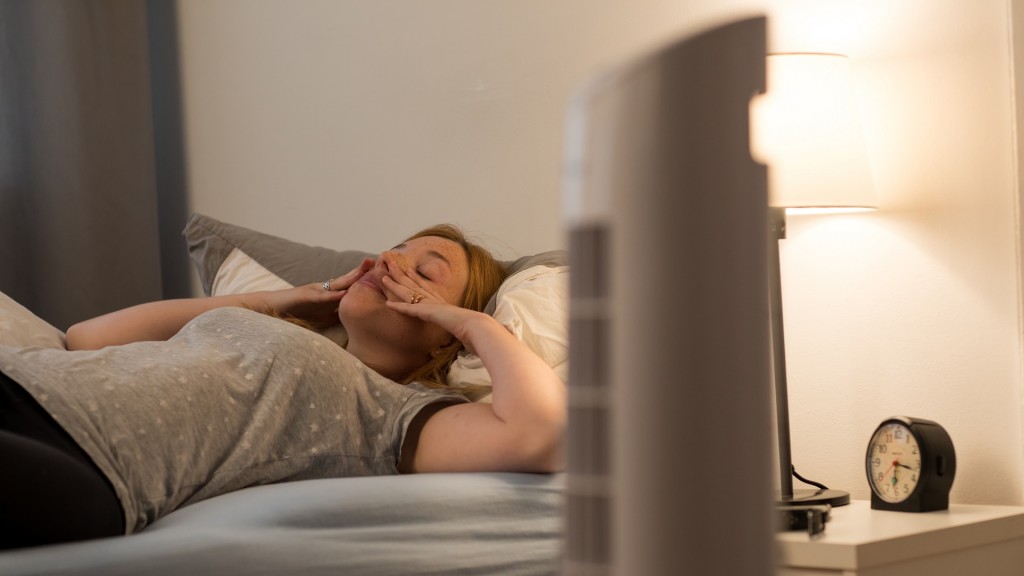 Eine junge Frau liegt im Bett, waehrend ein Ventilator davor steht und die Luft kühlt.