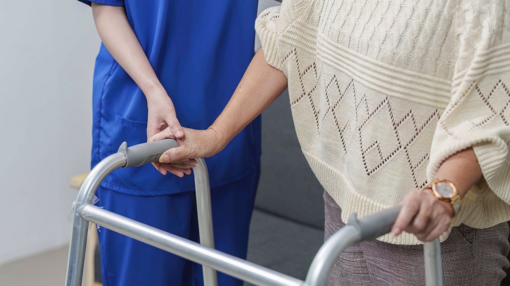 Eine Pflegekraft hält sanft die Hand einer älteren Person, die einen Rollator benutzt.