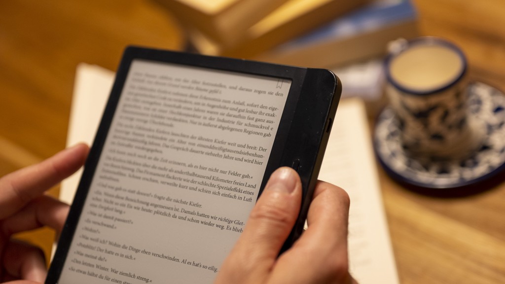 Ein Mann hält einen E-Book-Reader in der Hand, während im Hintergrund zahlreiche Papierbücher auf dem Tisch liegen.